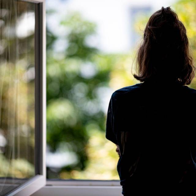 Eine junge Frau steht in ihrer Wohnung an einem Fenster