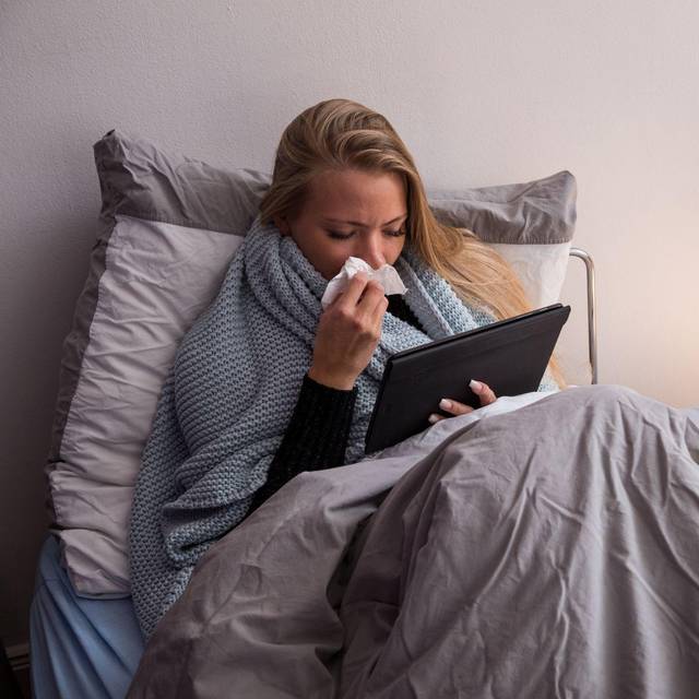 Eine Frau liegt krank im Bett und guckt auf ihren Tablet-Computer