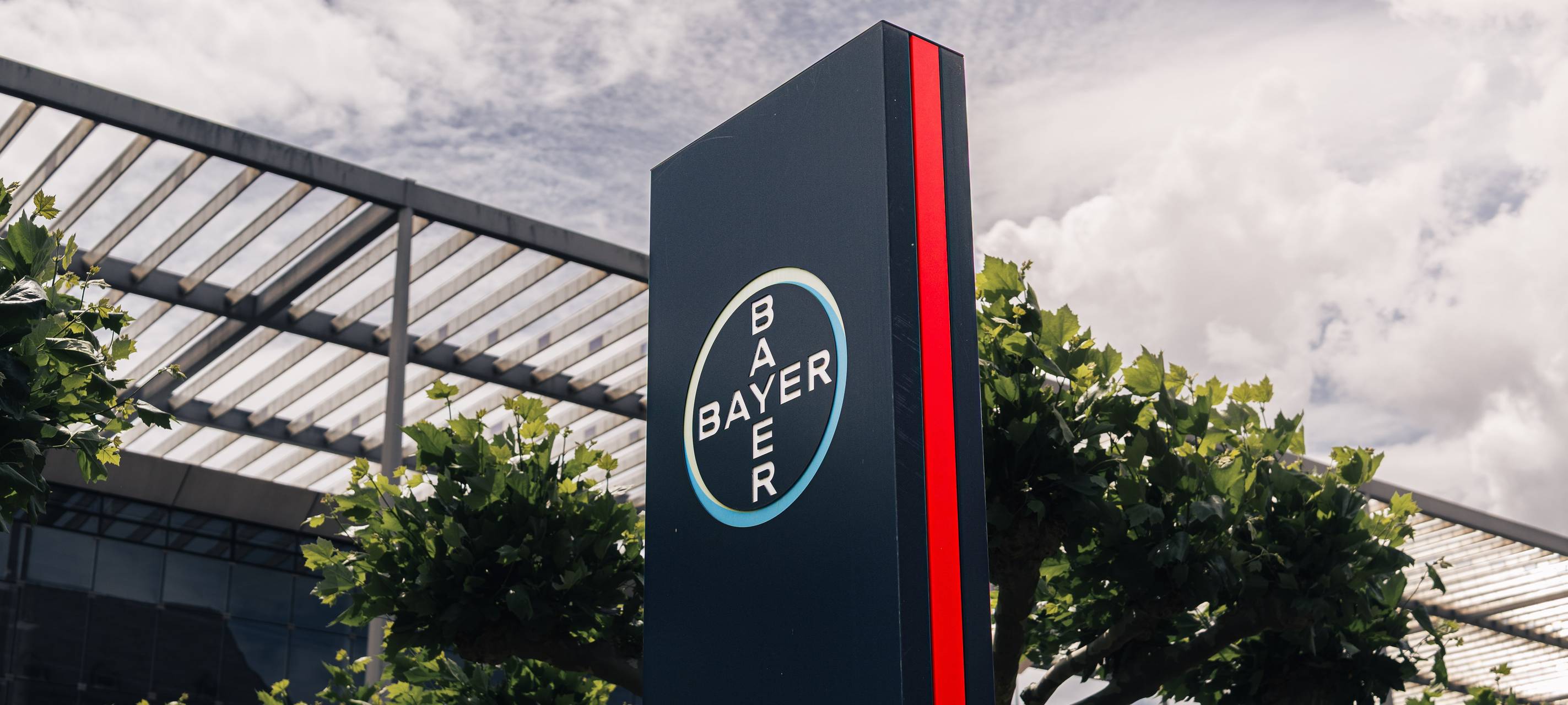 Stele mit Bayer-Logo und dem Schriftzug „Headquarters“. Im Hintergrund Bäume und Teile des Hauptquartiers von Bayer