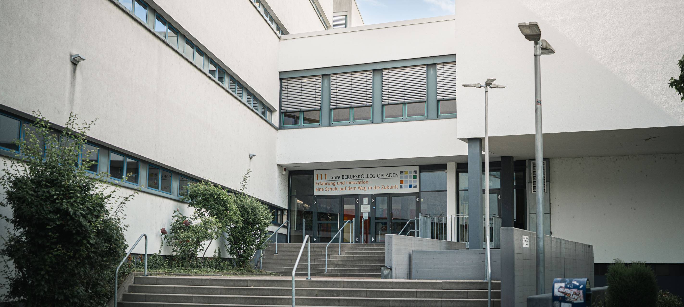 Eingangsbereich des Berufskollegs Opladen mit Treppenaufgang, Geländern und dem Schulgebäude im Hintergrund.