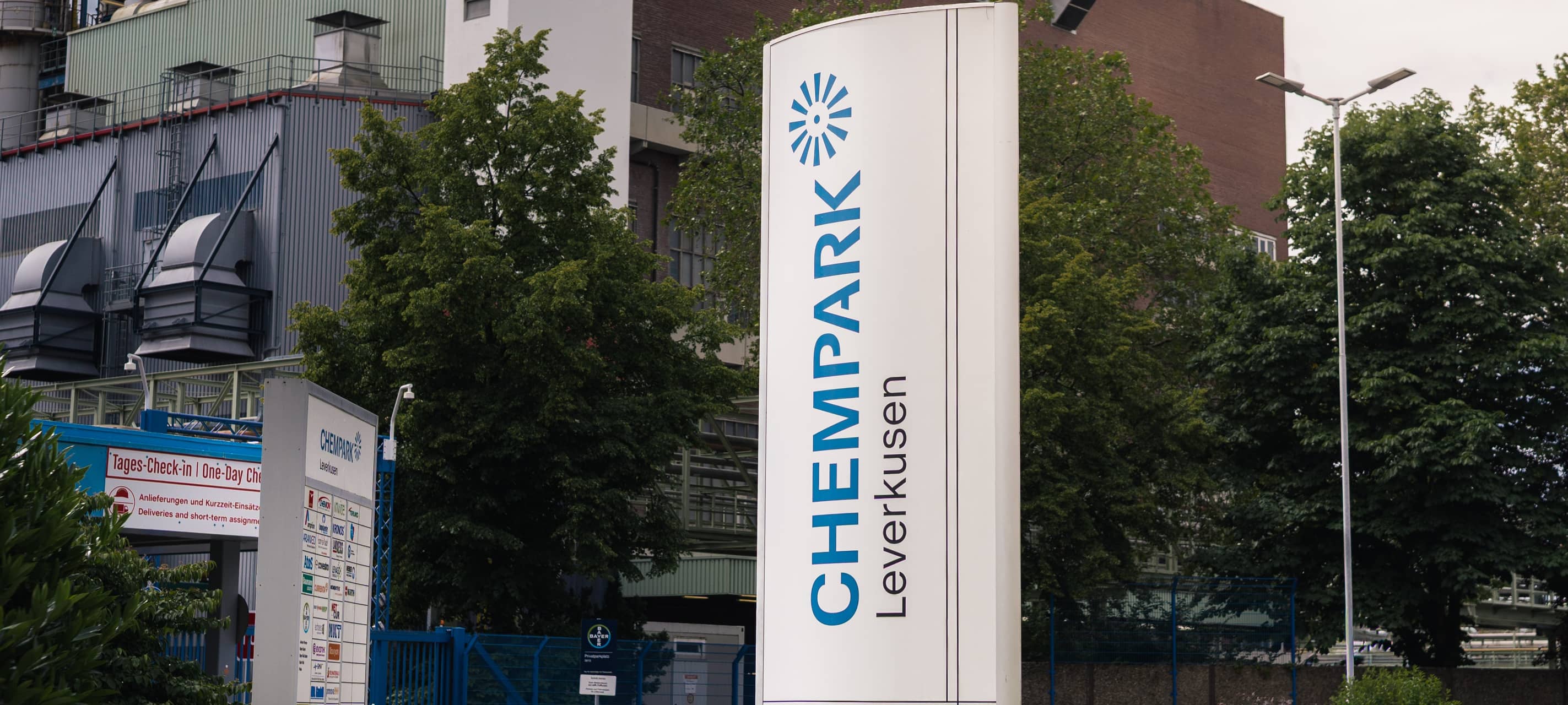 Stele mit der Aufschrift „Chempark Leverkusen“ vor Industrieanlagen und Bäumen.