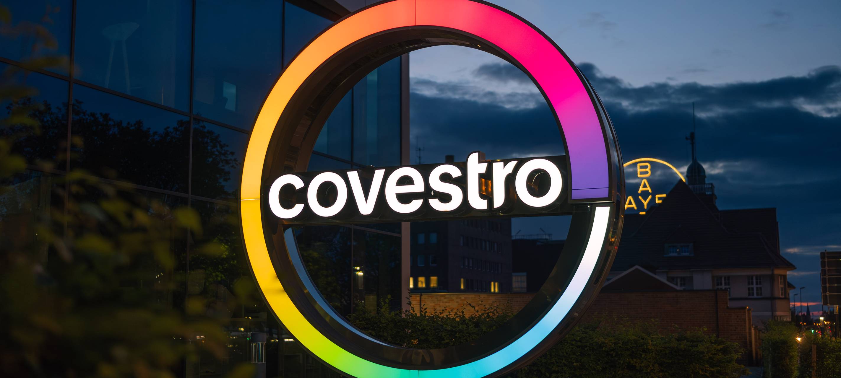 Beleuchtetes Covestro-Logo vor dem Hauptquartier in der Dämmerung; im Hintergrund ist das Bayer-Kreuz zu sehen.