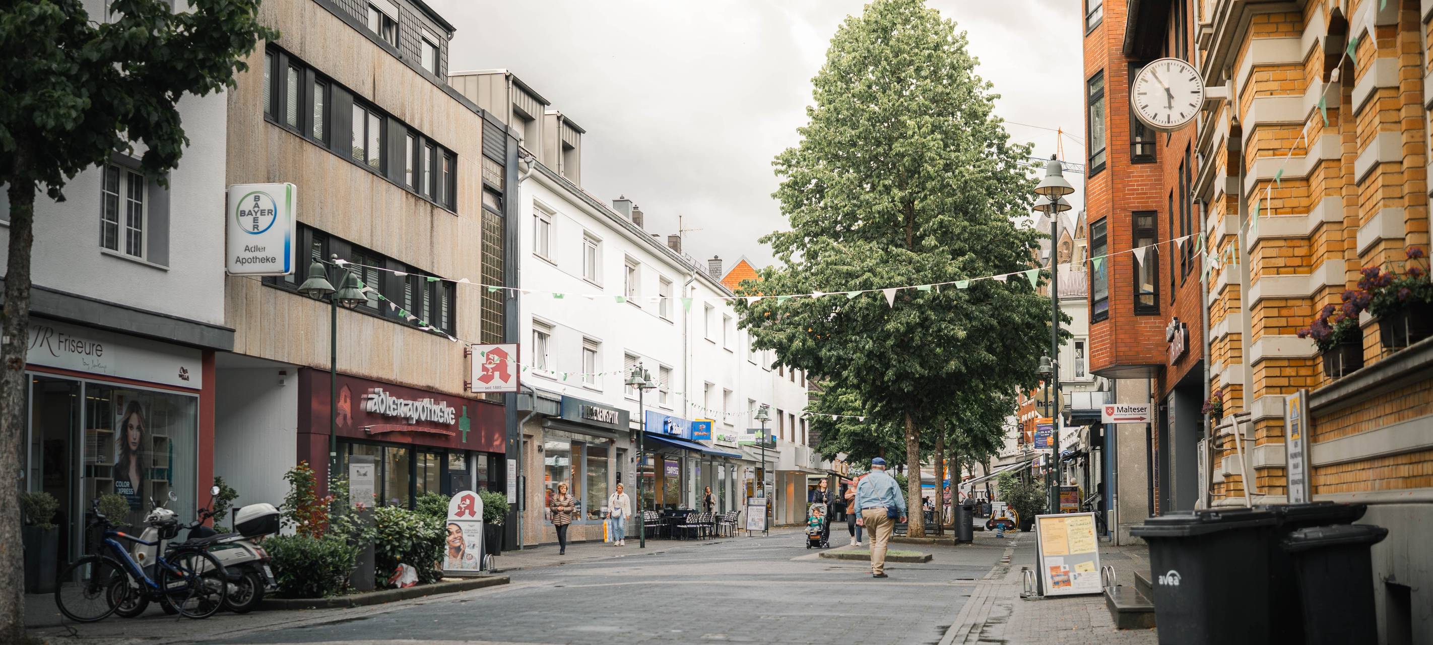 Fußgängerzone in Schlebusch mit Geschäften, Bäumen und einzelnen Passanten auf der Straße.