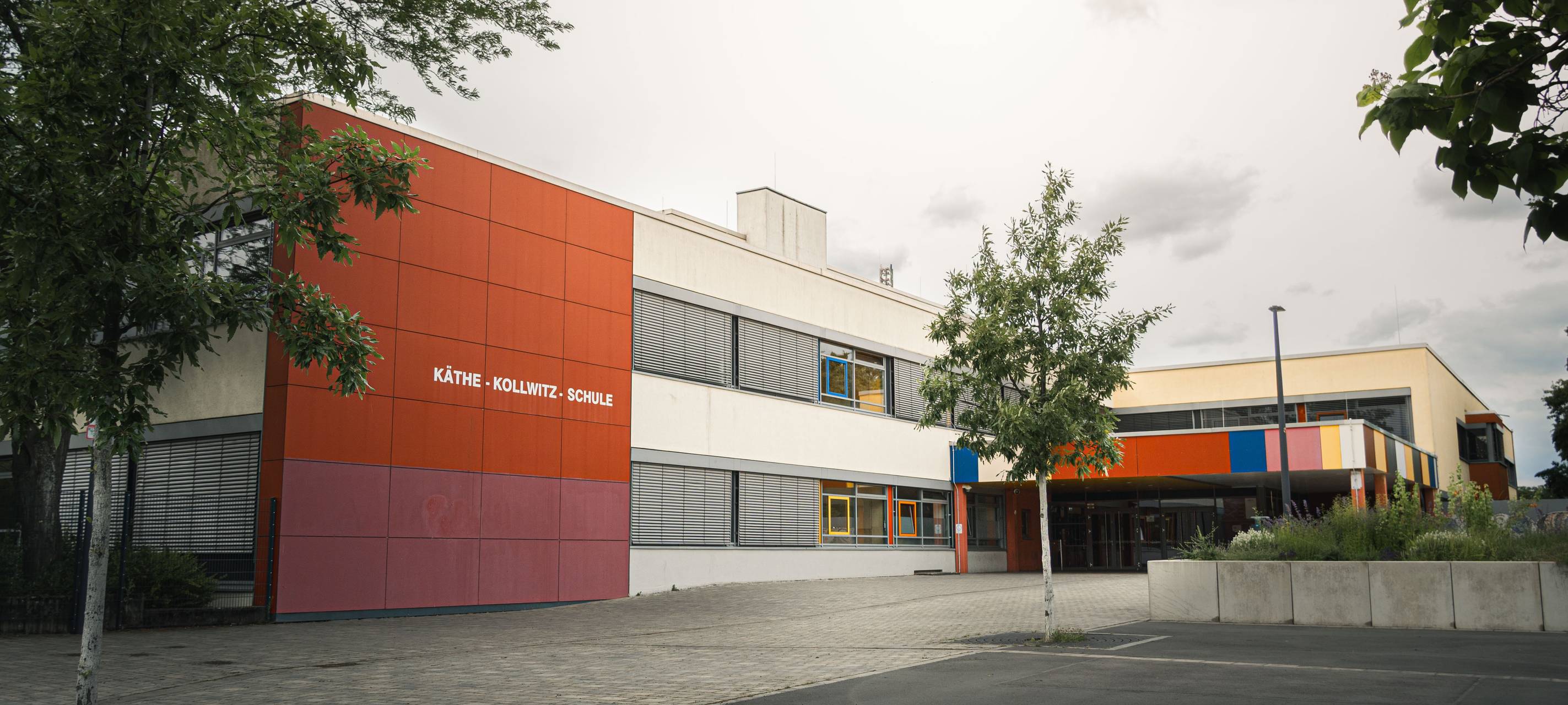 Gebäude der Käthe-Kollwitz-Schule mit farbiger Fassade, Vorplatz und Bäumen vor dem Eingang.