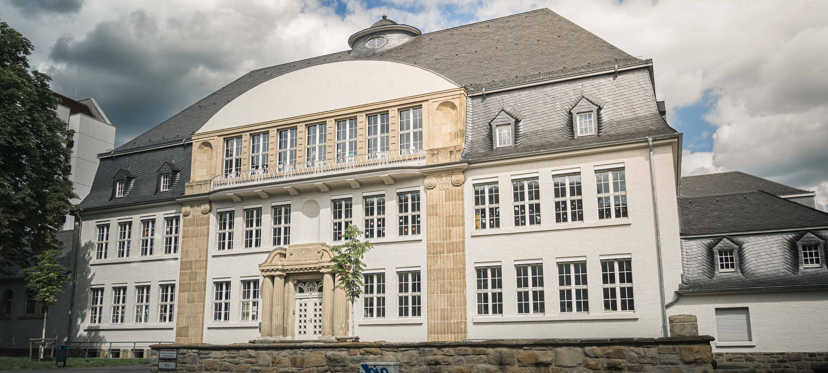 Schulgebäude der Katholischen Hauptschule Im Hederichsfeld mit heller Fassade und großem Dach.