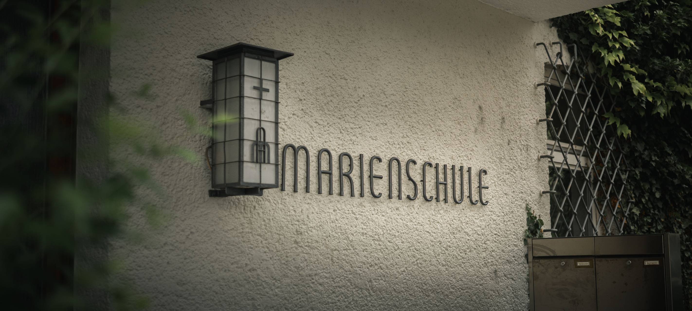 Schriftzug „Marienschule“ an einer hellen Hauswand neben einer Außenleuchte.